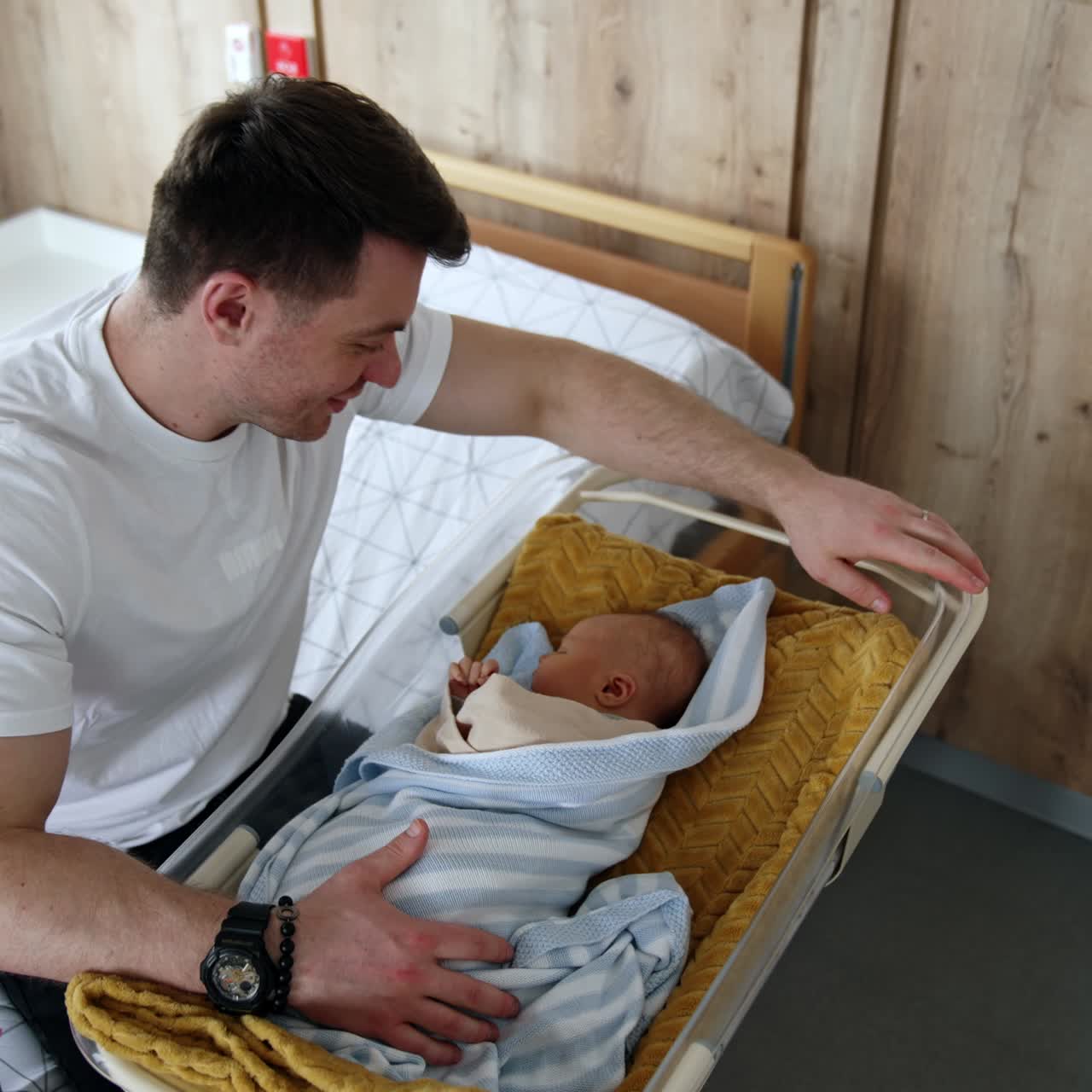 Loving dad sits at the crib with his baby. Happy father leans to kiss his newborn child on the head