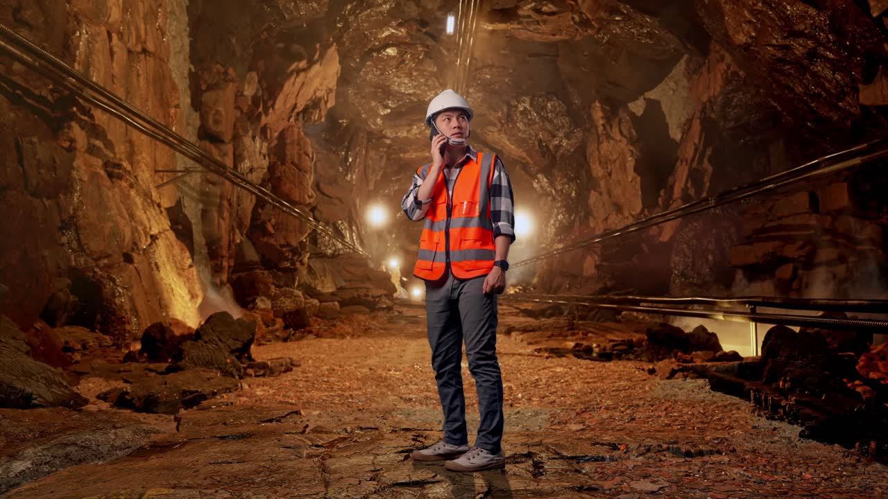 Full Body Side View Of Asian Male Engineer With Safety Helmet Talking On Smartphone While Standing In Underground Mine Tunnel