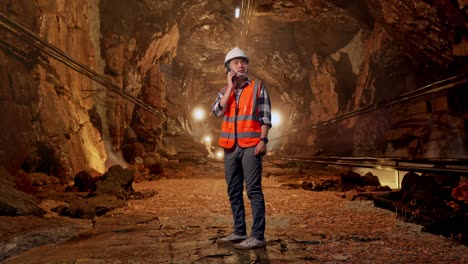 full body side view of asian male engineer with safety helmet talking on smartphone while standing in underground mine tunnel
