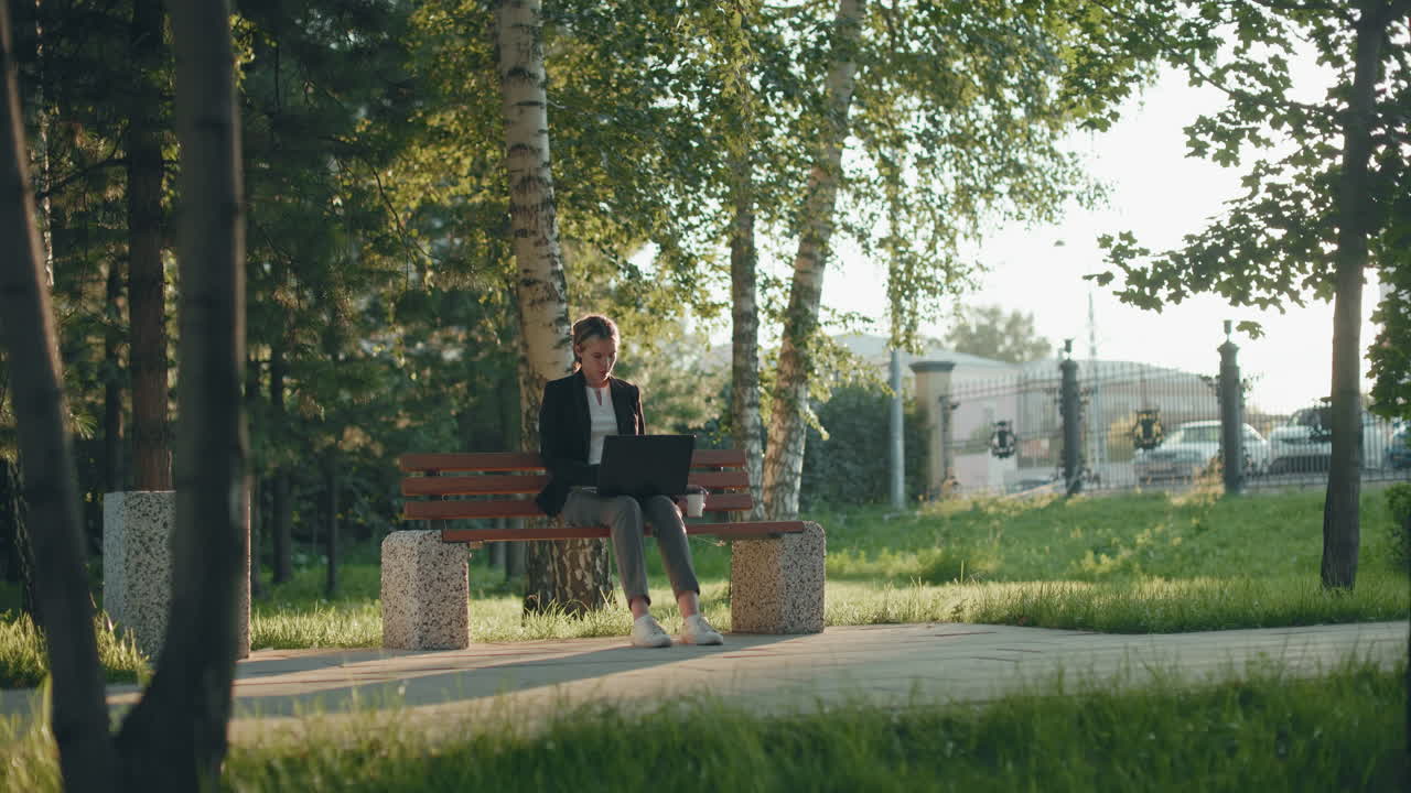 Young girl seated outdoor in lush park working on project with system while sipping fruit juice surrounded by trees, iron fence and parked car in background