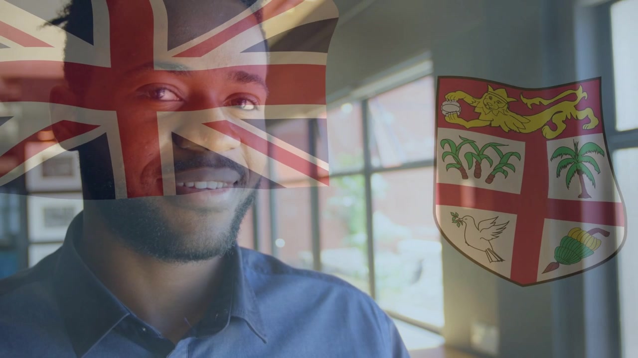 Mann står ved kontorvindu og ser lys, snur seg mot kamera og smiler, britisk flagg og skjold overlagt