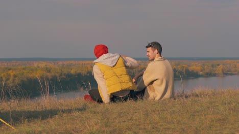 guy-in-vest-sits-down-on-river-bank-edge-and-hugs-boyfriend