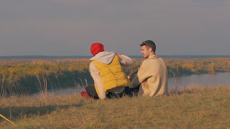 guy in vest sits down on river bank edge and hugs boyfriend