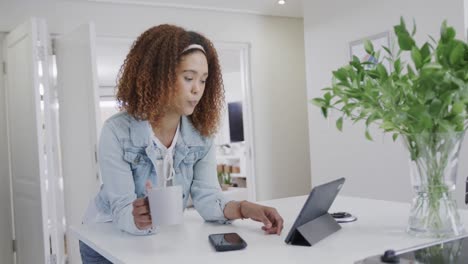 happy biracial woman using tablet and drinking coffee in kitchen, slow motion