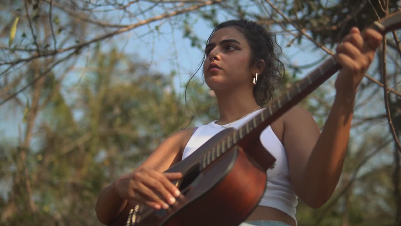 chica morena tocando una guitarra en un escenario con árboles y vegetación