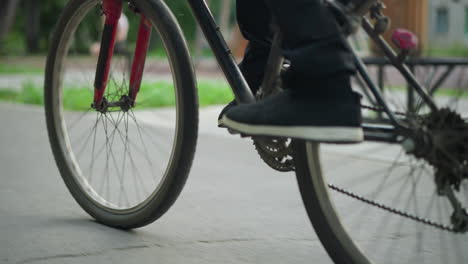 leg view of someone wearing sneakers riding a bicycle on paved road, captured from low angle, in the background, a blurred figure is sitting on a bench near greenery and trees