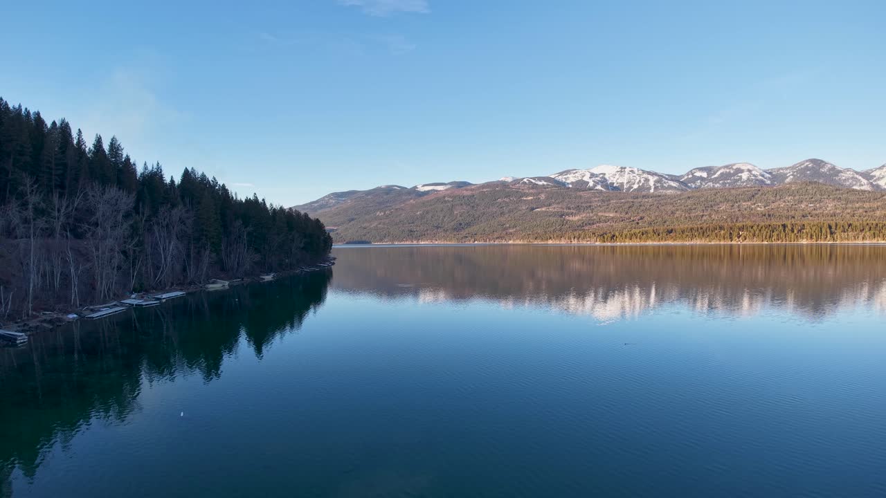 Whitefish, Montana yakınlarında, berrak mavi bir gökyüzü altında karla kaplı zirveleri ve her dem yeşil ormanları yansıtan sakin bir dağ gölü.