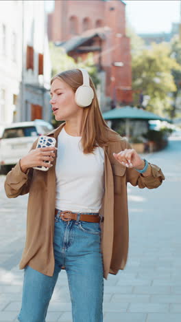 Happy woman in wireless headphones dancing listening favorite energetic music singing on street