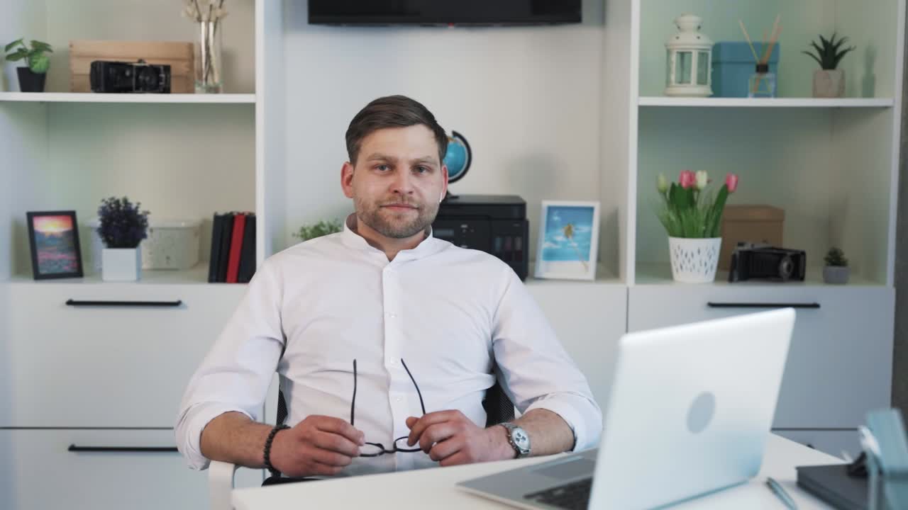Smiling adult man putting on and taking off eyeglasses with wireless headphones, wearing light shirt, standing against trendy light office background