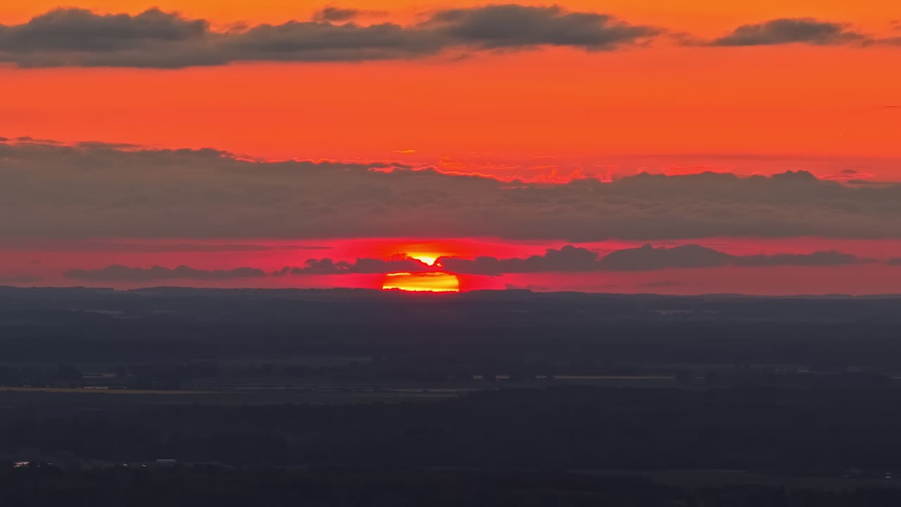 Sonnenuntergang hinter geschichteten Wolken über einer fernen Landschaft, tiefrotes und oranges Licht über den Horizont werfend. Atmosphärischer Abendhimmel mit glühender Sonne, die teilweise von Wolkenschichten verdeckt wird