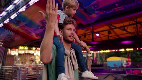 un padre feliz con el cabello rizado en una camiseta verde lleva a su pequeño hijo rubio en una camisa roja en sus hombros y miran a su alrededor en el parque de atracciones. luces brillantes e imágenes interesantes capturan el ojo e interés del pequeño niño rubio que se sienta en los hombros de su padre
