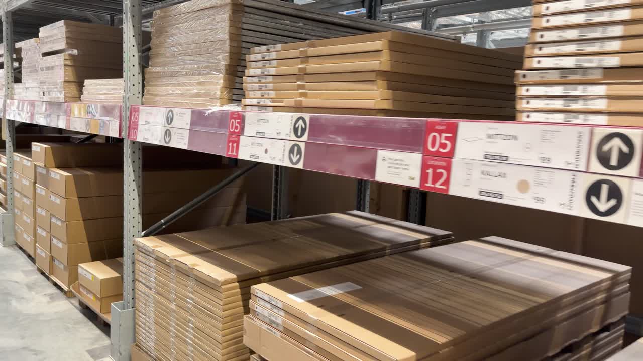 Camera moves along warehouse aisle stacked with flat-pack furniture boxes under bright industrial lighting