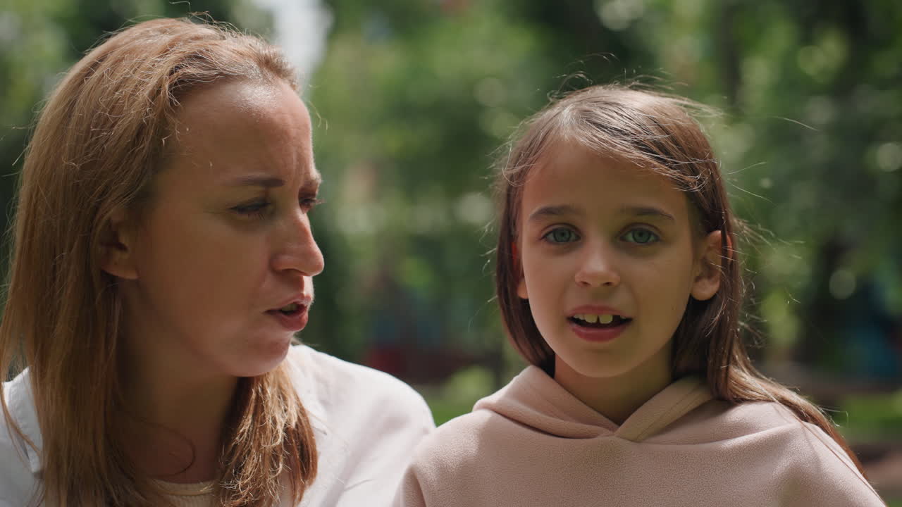 Mamma e figlia che sorseggiano una bibita al parco. La madre si china e scherza con la cannuccia mentre la bambina sorride e beve un sorso. Morbida luce solare filtra tra gli alberi, un primo piano spontaneo di un dolce legame e una conversazione tranquilla.