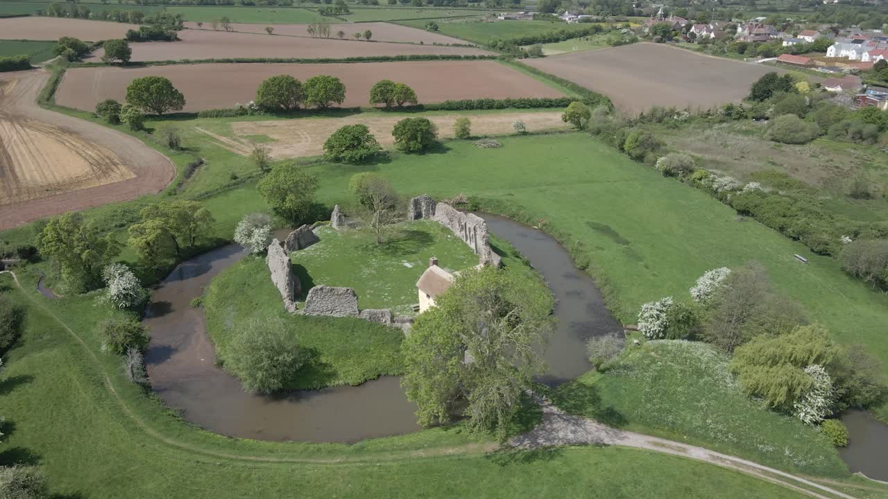 Aerial view of Stogursey Castle, a medieval castle in Somerset, England. Most of the site is in ruins, with a thatched gatehouse used for holiday's. Drone moving rotating over the ruins and moat.