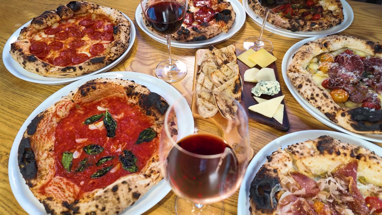 Draufsicht auf mehrere Pizzen, Rotwein und Vorspeisen auf einem hölzernen Restauranttisch.