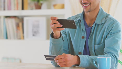 Hombre-En-Casa-Usando-Una-Aplicación-Bancaria-Tomando-Una-Fotografía-De-Una-Tarjeta-De-Crédito-Con-Un-Teléfono-Móvil