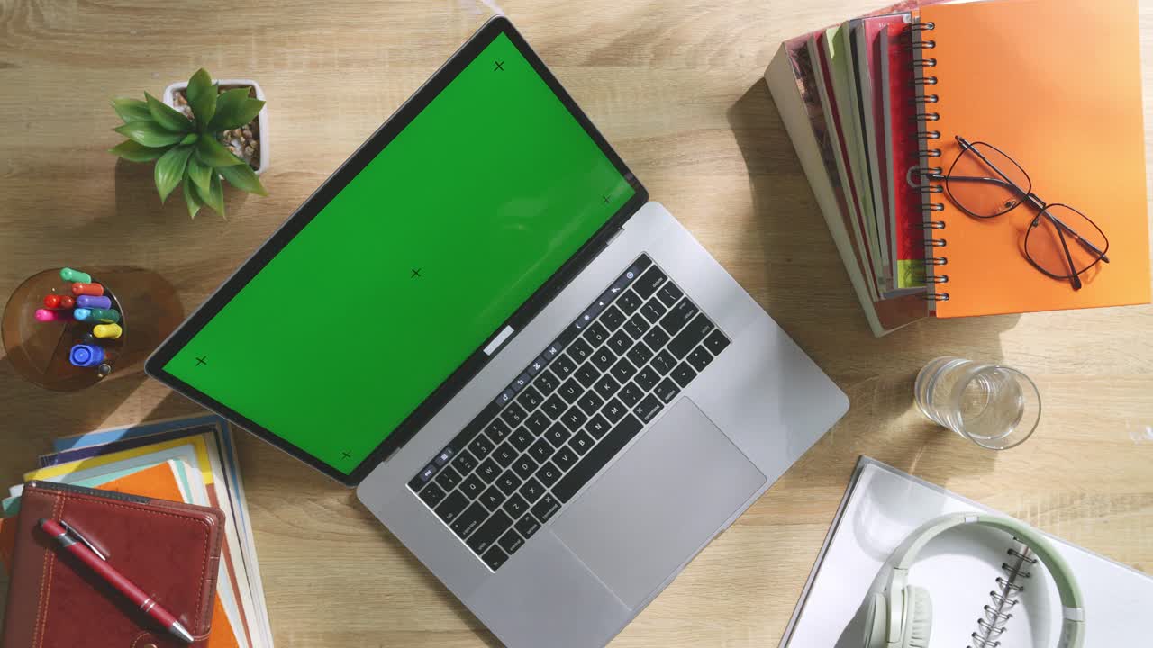 vista de arriba hacia abajo de una computadora portátil con pantalla verde simulada pantalla chromakey en un escritorio de oficina de madera al lado de la libreta con bolígrafos, gafas y un vaso de agua.