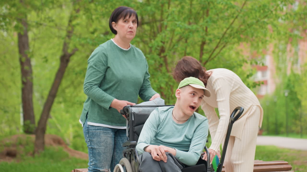 Park Scene White Man Wheelchair Caregiver Assisting, Middle Aged Mother Observes While Therapist Adjusts Harness, Green Cap, Bench By Path, Spring Trees, Serious Yet Compassionate Mood, Family Care