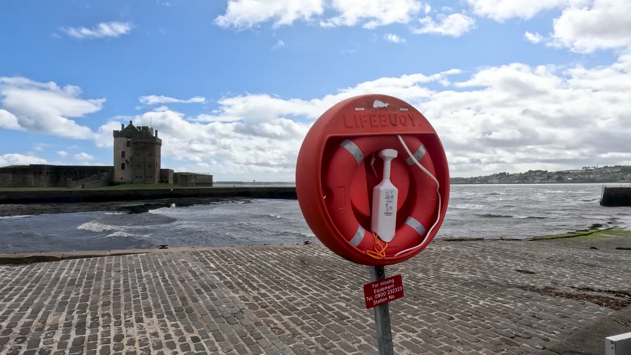 A red lifebuoy spins on its post near a waterfront, with Broughty Ferry Castle and choppy river under bright daylight and scattered clouds
