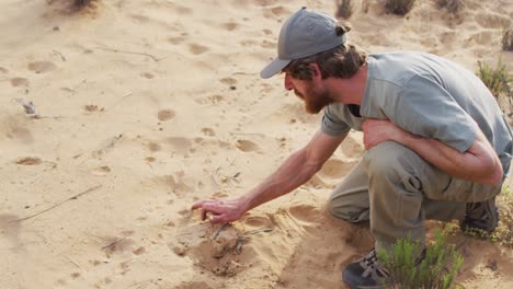 Caucasian-male-survivalist-in-wilderness-squatting-down-and-examining-animal-tracks-in-sand