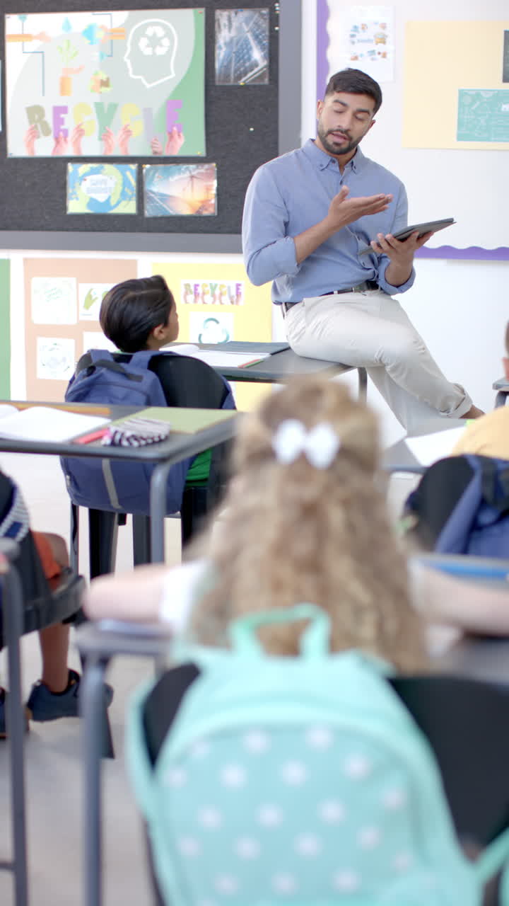 Asian male teacher with short black hair sits on desk, hand raised in vertical video
