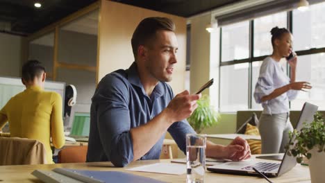 Happy-caucasian-businessman-sitting-at-table-and-using-smartphone-at-office