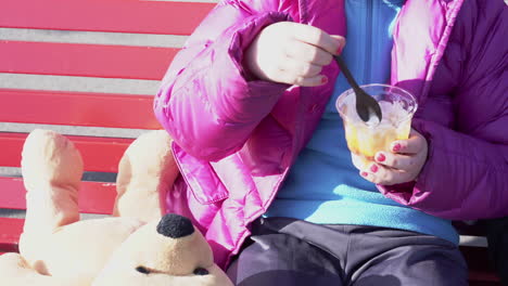 cerca de una niña con esmalte de uñas de puente azul y un oso de peluche a su lado sosteniendo un helado sundae sentado al aire libre 4k