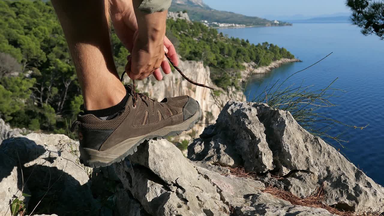 hombre excursionista en la cima de un acantilado de montaña con vista al mar atando cordones de zapatos, de cerca