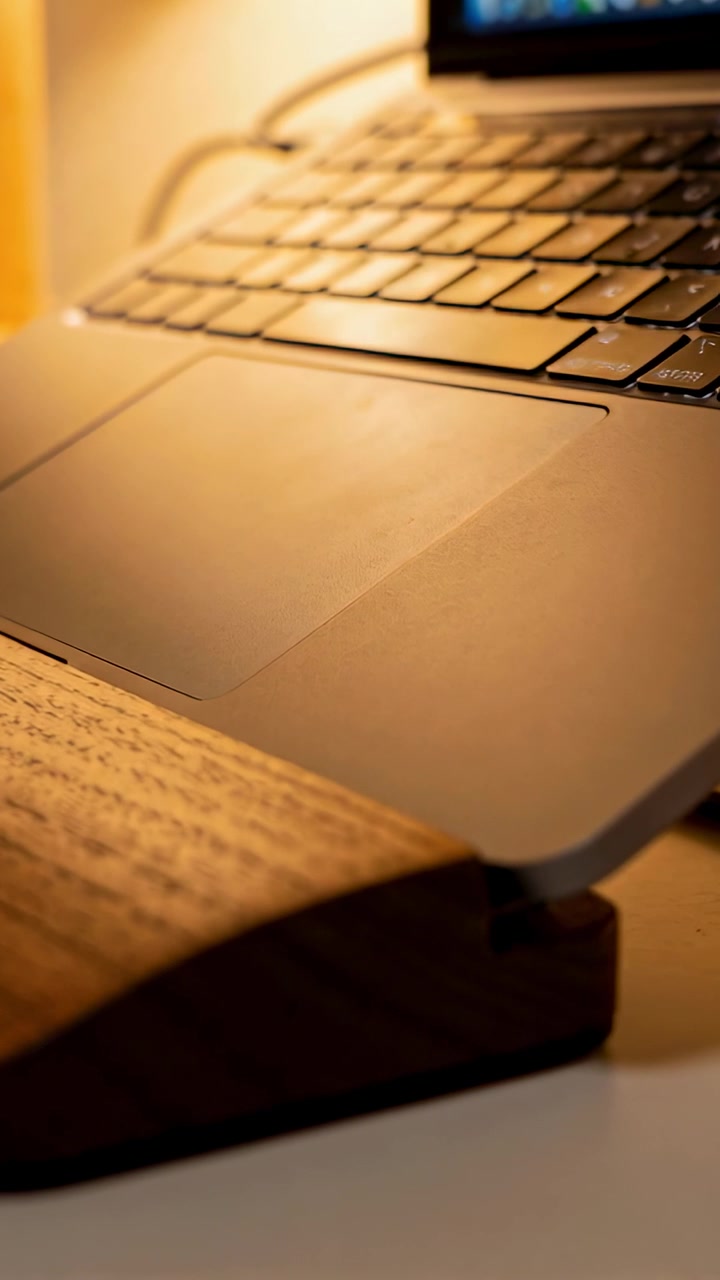 Vertical video: Tilting camera revealing aluminum laptop on wooden riser at desk, highlighting keys