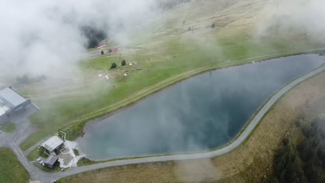 drone looks down on clouds floating over a beautiful small reservoir on the mountain asitz kogel in austria, leogang saalbach