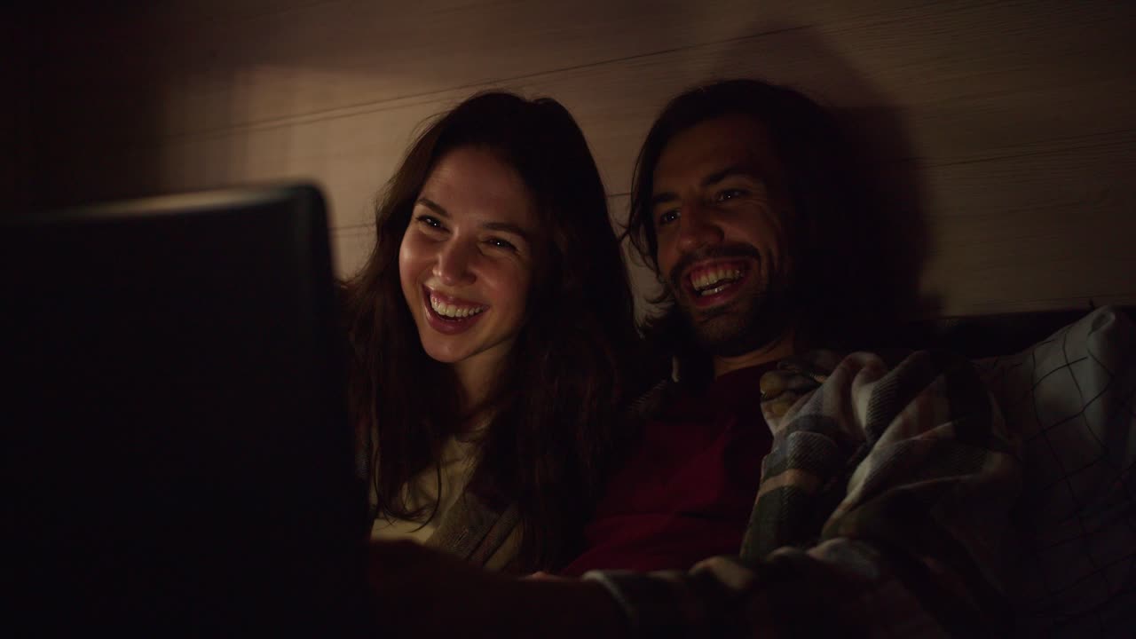 A happy couple, a brunette guy with stubble and his brunette girlfriend in a plaid shirt, lie on the sofa in a trailer and watch a funny comedy using a laptop with dimmed lights in an atmospheric romantic evening near a camp in a trailer outside the city in the summer