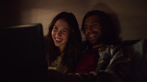 A-happy-couple,-a-brunette-guy-with-stubble-and-his-brunette-girlfriend-in-a-plaid-shirt,-lie-on-the-sofa-in-a-trailer-and-watch-a-funny-comedy-using-a-laptop-with-dimmed-lights-in-an-atmospheric-romantic-evening-near-a-camp-in-a-trailer-outside-the-city-in-the-summer