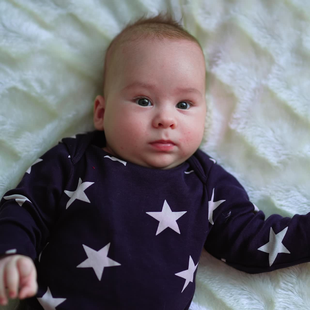 En sunn og aktiv spedbarnsgutt fryser et øyeblikk og ser rett frem. Vakker baby som smiler lett mens han ligger på en hvit seng. Nærbilde