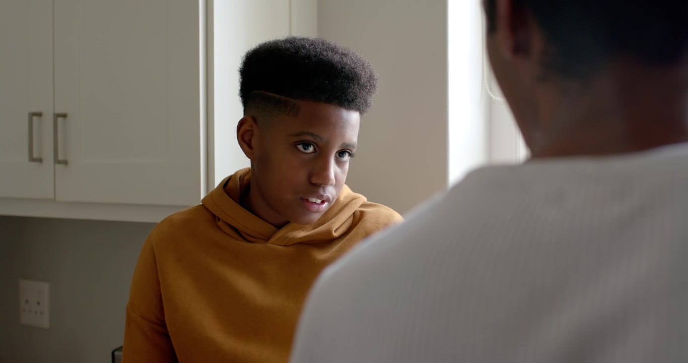 Teen male in mustard hoodie looking up softening smile listening at home while adult speaking right