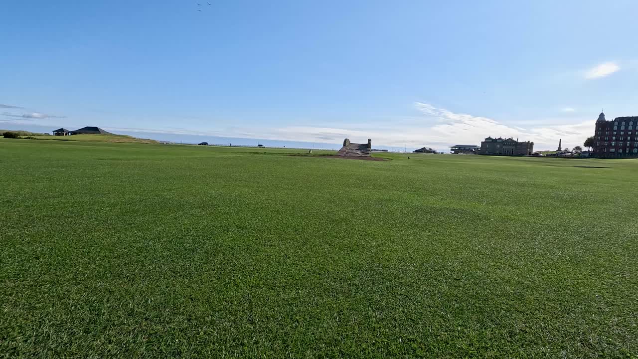 Wide-angle pan over green golf course, historic buildings, blue sky, bright natural daylight