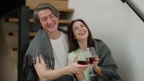 Happy-couple-sit-on-the-wooden-stairs-hold-glasses-drink-red-wine-talk-laugh-hugging