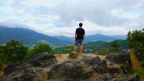 man-walking-down-from-a-lookout-over-a-valley,-houses-climbing-up-the-side-of-mountains-sprawling-forests-climbing-up-the-hills-to-cloud-covered-tips