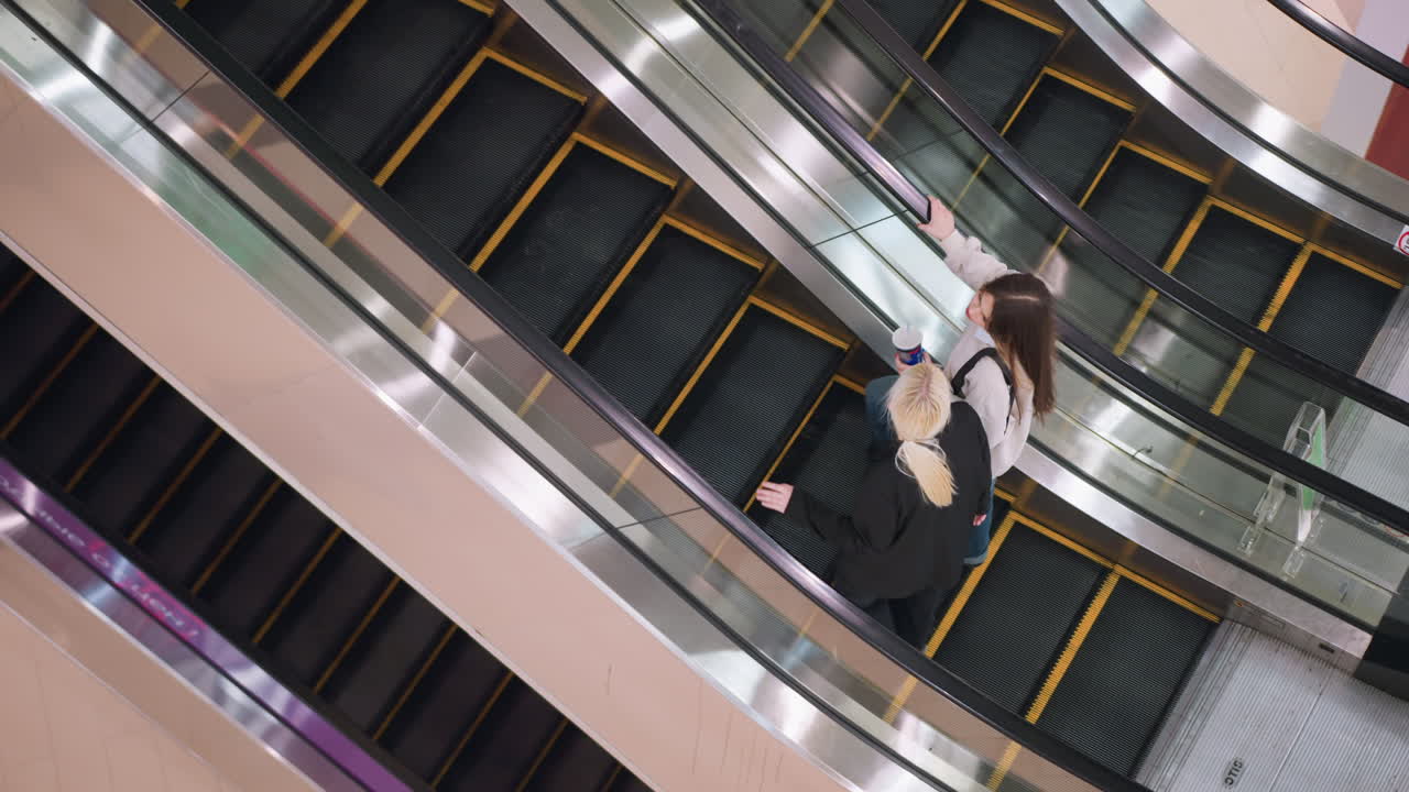 Overhead view of two ladies stepping on ascending escalator inside shopping mall, talking casually while one holds drink, capturing relaxed lifestyle moment and social interaction