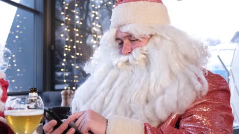 Santa Claus Uses a Smartphone while Sitting in a Cafe he is Resting and Talking on the Phone Santa Claus Uses a Smartphone while Sitting in a Cafe he is Resting and Talking on the Phone Funny Santa Claus