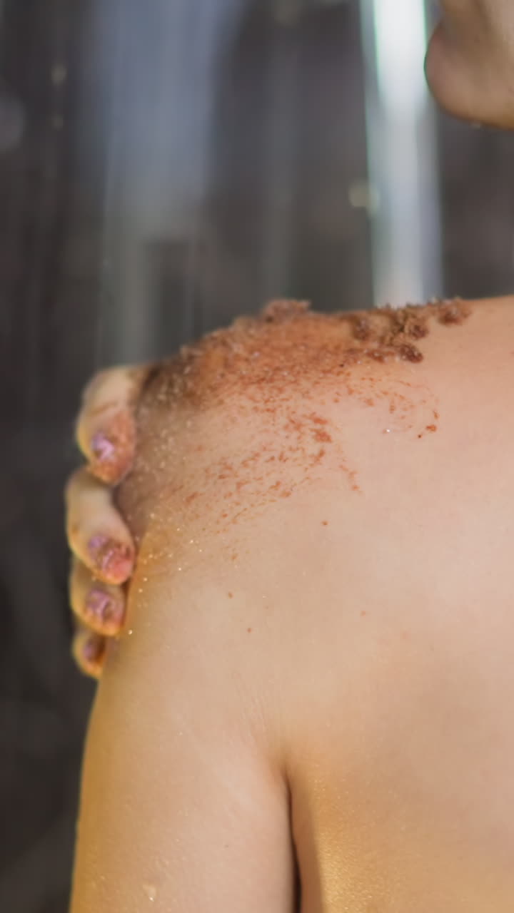 Young woman takes shower spreading dark coffee scrub on bare shoulder in circular motion with palm in bathroom skincare daily routine closeup slow motion
