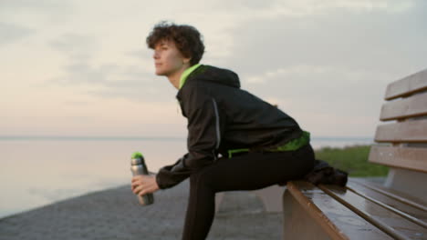 young woman in sportswear sitting on bench on riverside promenade, holding water bottle and watching beautiful sunrise before morning run