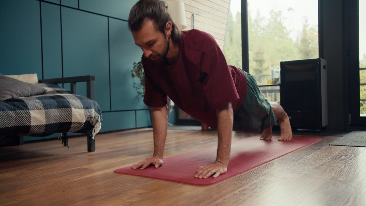 un tipo moreno con una camiseta roja hace flexiones en una alfombra especial en una casa de campo con vistas a un bosque verde