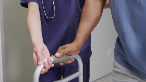 midsection of diverse female doctor helping male patient use walking frame at hospital