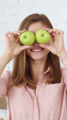 the girl in the white kitchen is having fun with green apples, closing her eyes and smiling. vertical video