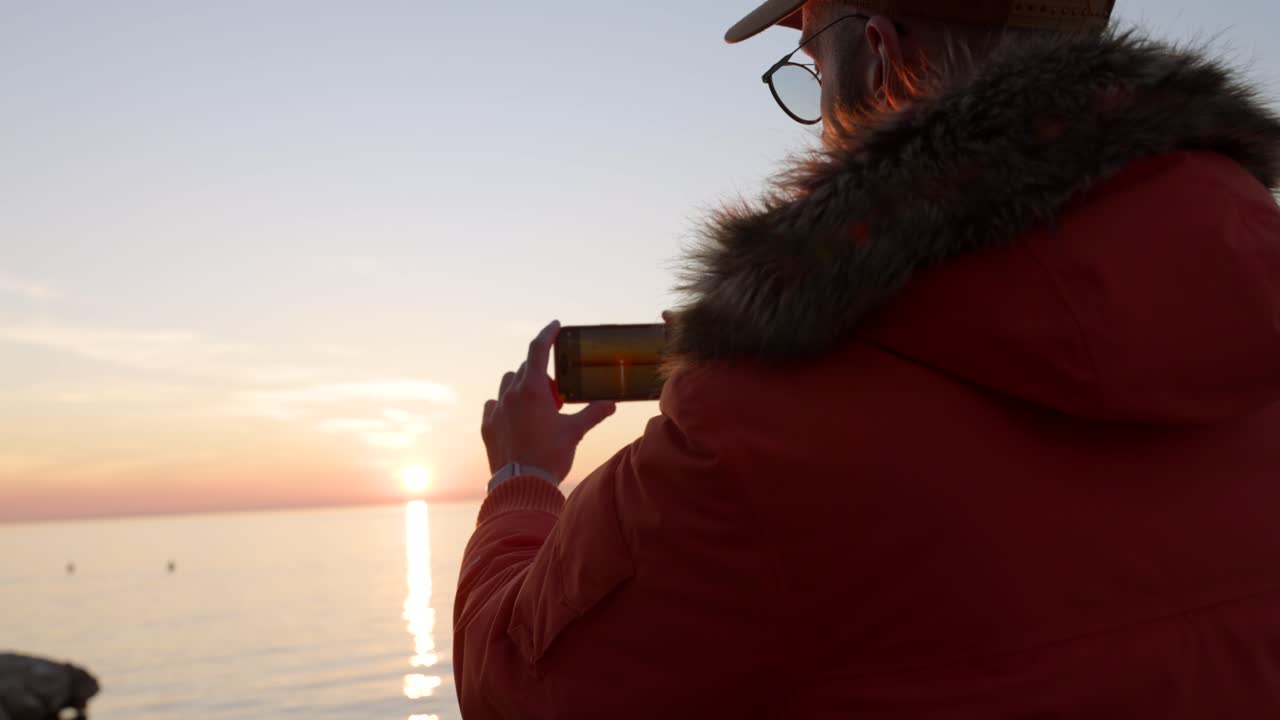 hombre con una chaqueta naranja tomando fotos con su smartphone de la puesta de sol detrás del océano