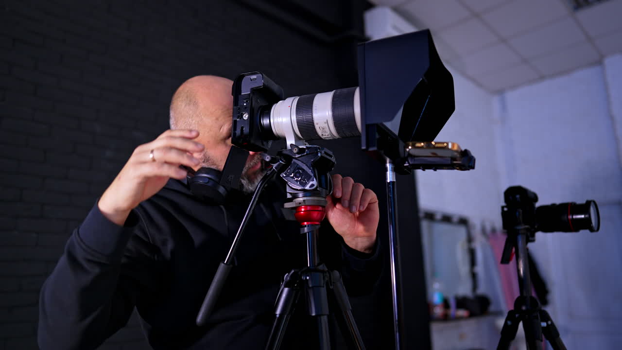 Videomaker che regola una telecamera professionale con teleobiettivo su treppiede. Un uomo utilizza una telecamera digitale di fascia alta con obiettivo bianco in un ambiente studio con attrezzatura di illuminazione e monitor.
