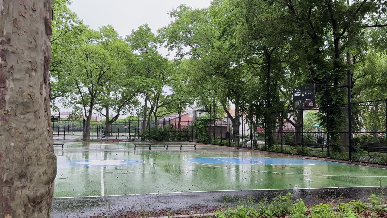 Static shot of a wet basketball court in Bushwick, framed by trees and a passerby in the background.