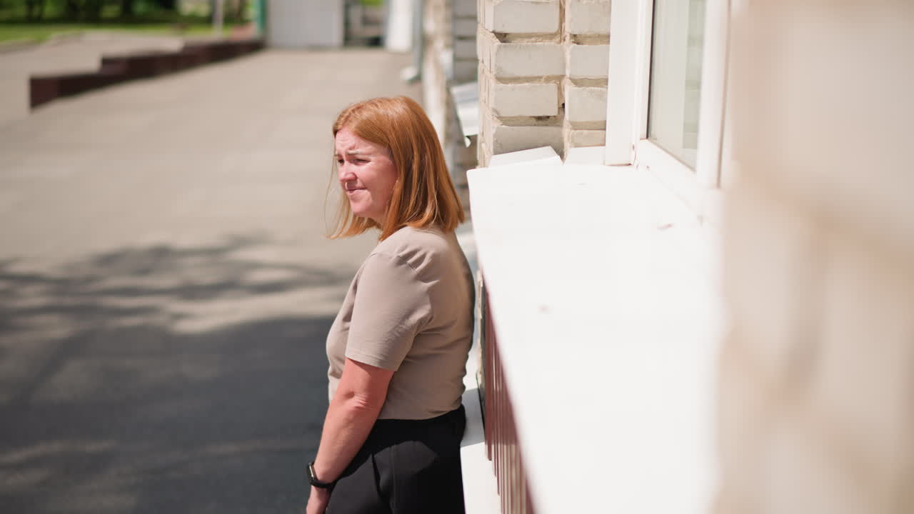 Tired woman walking toward brick building under warm summer sunlight, holding bag and pausing beside wall, expressing exhaustion, quiet reflection, and calm moment in peaceful
