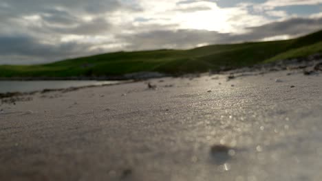 Eine-Aufnahme-Von-Goldenen-Sandkörnern-An-Einem-Strand-Aus-Einem-Niedrigen-Winkel,-Während-Die-Sonne-Hinter-Grasbewachsenen-Hügeln-Und-Tosenden-Wellen-Im-Hintergrund-Untergeht