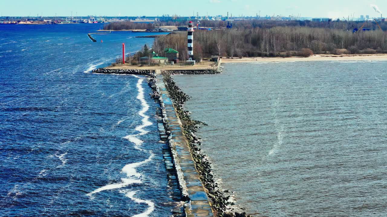 A bold black-and-white lighthouse stands past a rugged stone pier, where vivid blue water meets pale sand and leafless trees under a clear sky.